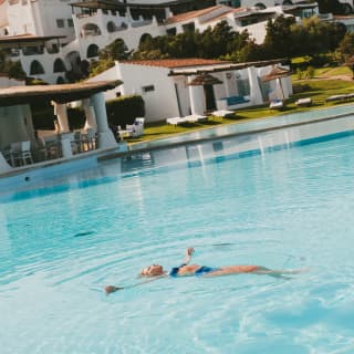 A woman in a blue bikini floats on her back in the otherwise empty crystal pool in front of lawns and lounge areas.