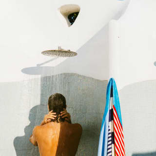 A man takes a shower in an outside cubicle below a blue sky with his blue, red and white towel on a peg, seen from behind.