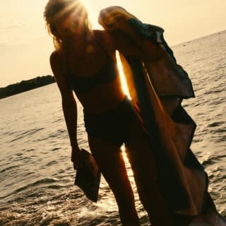 Angled image of woman in the shallow white waves, holding a magazine and a towel, silhouetted against the golden sunset.