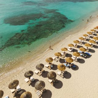 Two rows of parasols and sun-beds follow the sandy curve of the bay where people wade in the emerald waters, seen from above.