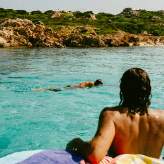 A man sits on a boat's diving platform, back against the towelled deck, as his friend swims in the sea, seen from behind.