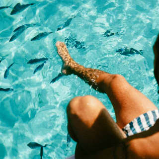 A man in blue stripy trunks swishes his leg in the incredible emerald waters, teeming with fish, seen from above.