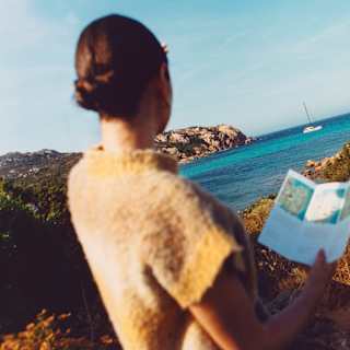 In soft-focus foreground, seen from behind, a woman holds a hiking guide and gazes at an azure bay along the Pevero Trail.