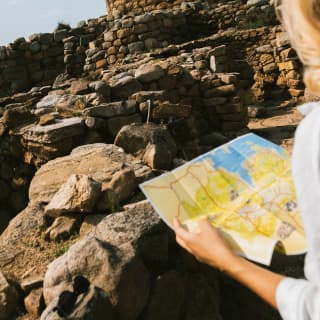 A circular ancient nuraghe fortress made with stone walls, seen from over the shoulder of a tourist regarding a map.