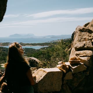 Sitting on a stone wall in the hills, a woman gazes over forest towards Pevero Beach and islands beyond, viewed from behind.