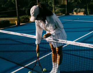 A woman in tennis whites and cap leans over the net with her racket, where two yellow balls rest on the blue surface court.