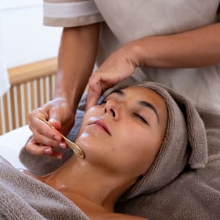A facialist uses a sculpting tool on a female guest with head and chest wrapped in taupe towels during a collagen treatment.