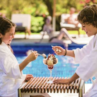A boy and girl in white towel robes sit either side of a poolside table sharing an ice-cream in a glass with chocolate sauce.