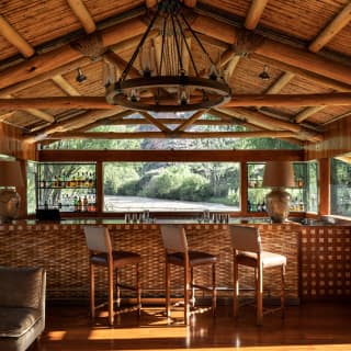 Beneath a wood-beamed roof, three high-back stools sit along the bar at El Huerto Bar with a direct view to the Urubamba River.
