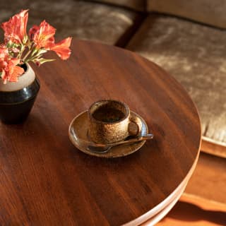 A stunning gold-tone stoneware cup and saucer, with spoon, rests on a round wood table with a vase of orange Peruvian lilies.