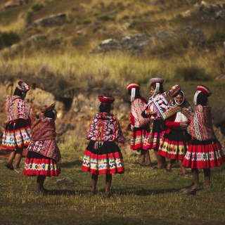 Women gather on a grassed area, dressed in vibrant traditional skirts, Manta shawls and Montera hats with sanq'apa straps.