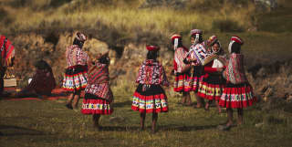 Women gather on a grassed area, dressed in vibrant traditional skirts, Manta shawls and Montera hats with sanq'apa straps.
