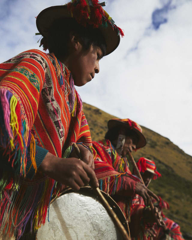 A low-angle shot of a man in a woven Manta as he plays a cumaru wood drum alongside other musicians celebrating Pachamama.