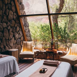 A window on to untamed countryside lets dappled light into a mellow spa treatment room with two beds, two chairs and candles.