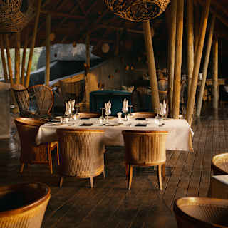 A wicker pendant lamp hangs in the restaurant, complementing wood beams and chairs, seen around a laid white-clothed table.