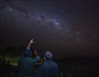 A group of star-gazers, barely visible in the dark, point at the glittery smudge of the milky way in the night sky above.