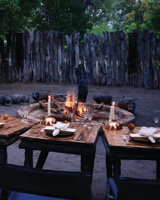 Candlelit dining tables arching around a circular campfire in a shaded clearing