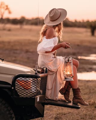 Stylish lady in a flowing white dress holding a lantern and perching on a truck bonnet