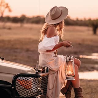 Stylish lady in a flowing white dress holding a lantern and perching on a truck bonnet