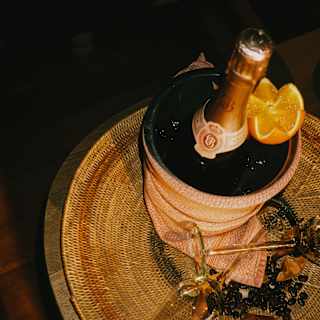 A bottle of Champagne cools in an ice-bucket, presented with an orange-half on a wicker mat with two flutes, seen from above.
