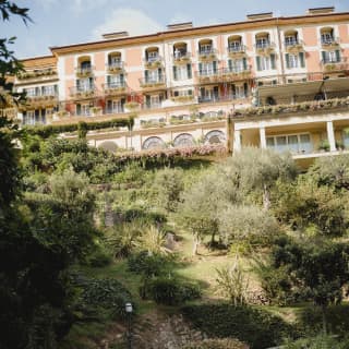 Two women walk in along a shady path below the palatial hotel, which sits atop the hill in a splendour of blushing hues.