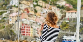 A woman holds onto a part of the rigging aboard a sailing boat in the bay. Before her pastel coloured houses climb the cliff