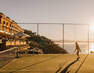 A woman, half-silhouetted against the blaze of the sun, plays tennis on a court overlooking the sea and the hotel and gardens.