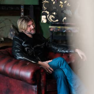 The fresco artist Gio Bressana sits in a Chesterfield style leather armchair, surrounded by ornate, gold leaf Chinoiserie