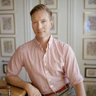 Acclaimed interiors architect and designer, Martin Brudnizki, leans on a table edge. Behind him, a wall of gilt framed pictures