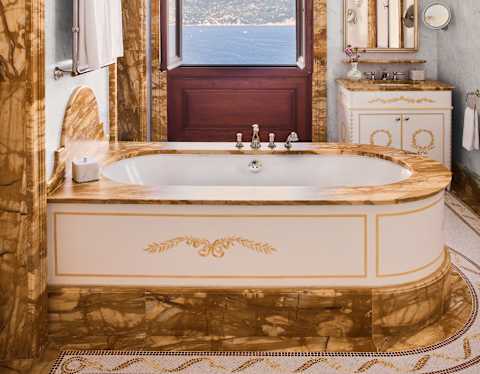 Looking through a white bathroom with ochre marble details, in door fames and a luxury bath, with an open, sea-view window.