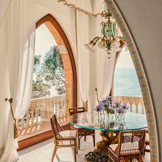 A glass-topped dining table with allium bud vases bathes in light and views from wide arched windows on perpendicular walls.