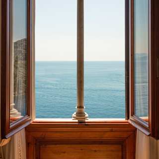 An open double window, with a wooden frame and a slim stone central support, offers breathtaking ocean views and blues.