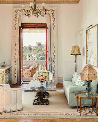 A tall, open door brings sublime views into an elegant room in pastels, with a chandelier, grey sofa and marble coffee table.