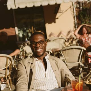 A young man in cream linen blazer smiles at the camera from a bar terrace. A glass of Negroni is on the table in front of him