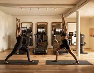Two women in black yoga wear hold the warrior one pose as they face each other on separate mats, seen in a wood-floored gym.