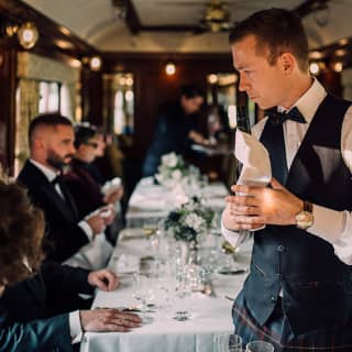 Preparing for dinner, a waiter in black and tartan holds a wine bottle in a cloth as guests settle at immaculately-set tables.