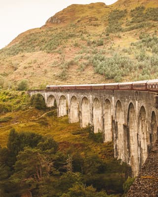 Looking from a window to the front of the train, which pulls carriages over the iconic Glenfinnan Viaduct towards a hillside.