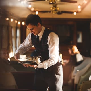 Steward in a tartan waistcoat holding a silver tray with a teacup and sugar bowl
