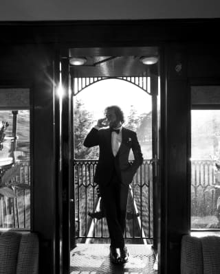 In black and white, a male guest in black-tie sips a drink with one hand in his pocket on the open-air observation deck.