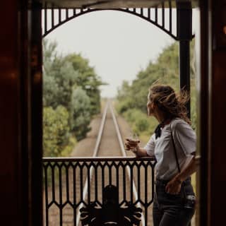 Wind flicks the long hair of a woman as she rests on the deck railings with a drink and watches the trailing landscape.