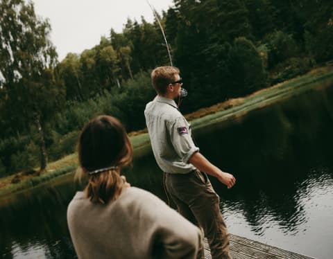 At a lake's edge, a man casts a fishing rod from a wooden deck watched by a woman with her hand on her hip, seen from behind.