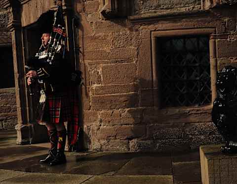 Eye-level close-up of Glamis Castle with a man in traditional costume playing bagpipes, left, and a black lion statue, right.