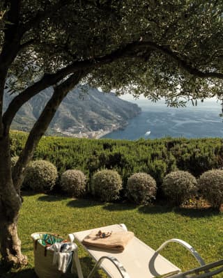 In the tranquil gardens, a basket and a sun lounger wait for a guest beneath the boughs of an olive tree, facing the coast.