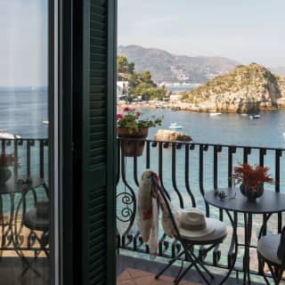 A panama hat lies discarded on a private balcony chair. The view stretches across the Bay of Mazzarò to a white rocky outcrop