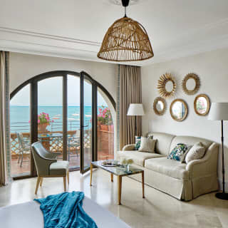 Small boats with canopies bob on the sea. Arched glass doors give access to the private balcony from an elegant sitting room