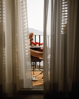 View from a shadowy suite through diaphanous drapes onto the balcony where a guest sits with feet up, enjoying the views.