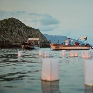 White paper box lanterns float on the calm evening sea as a young couple in a boat clink glasses on their romantic getaway