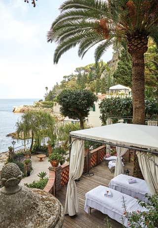 Massage tables are set up with white towels within a curtained cabana on a garden terrace, surrounded by trees and flowers>