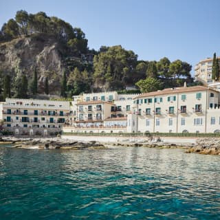 View from across the water of a hotel with a cream facade and clay tiled roof