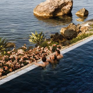 Three swimmers rest at the edge of the pool, gazing out past a small rock garden beach and nearby sea stacks to the ocean.
